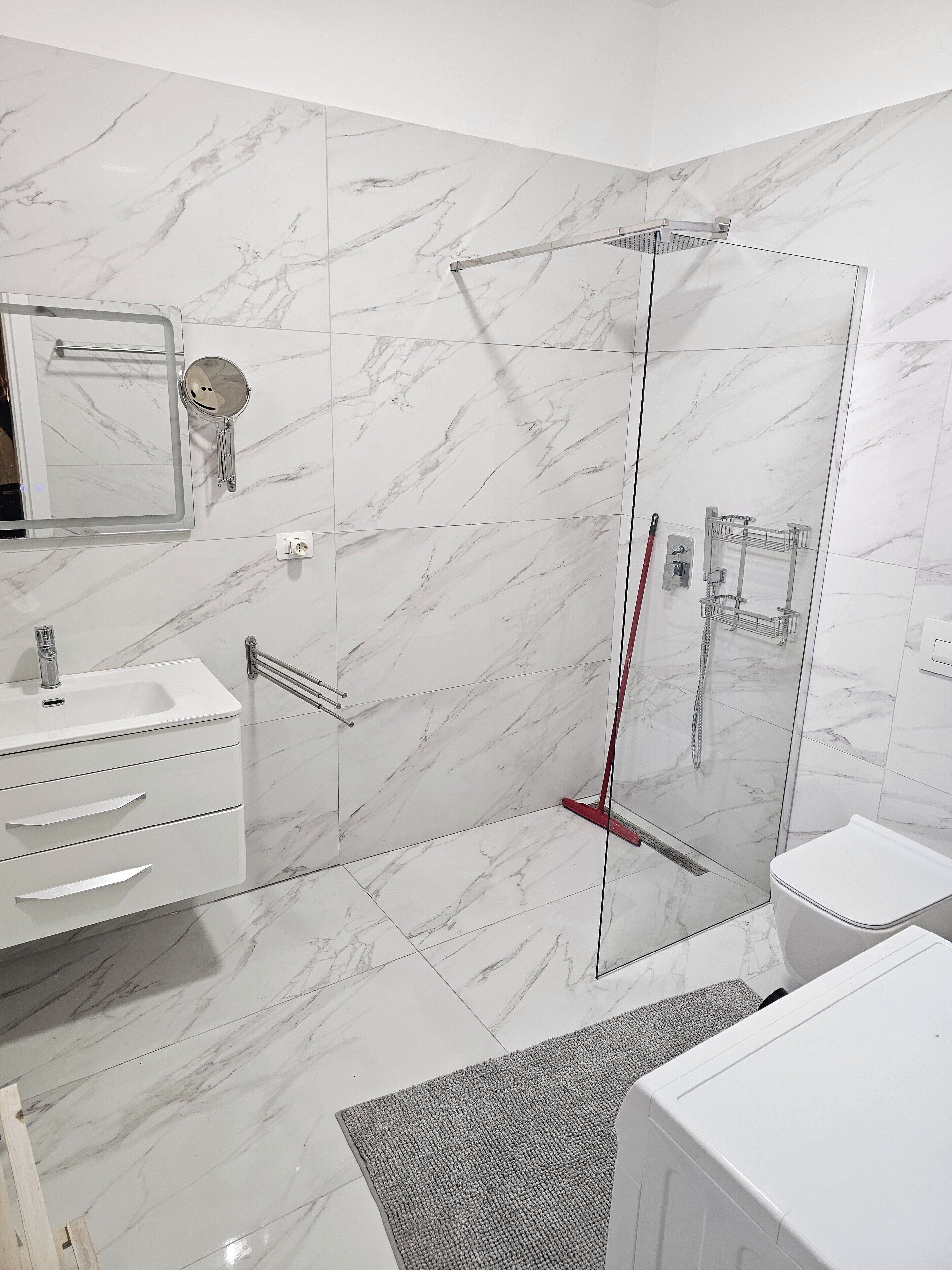 A modern bathroom with a glass shower enclosure, white marble tiles, a white sink with a mirror, and a white toilet.