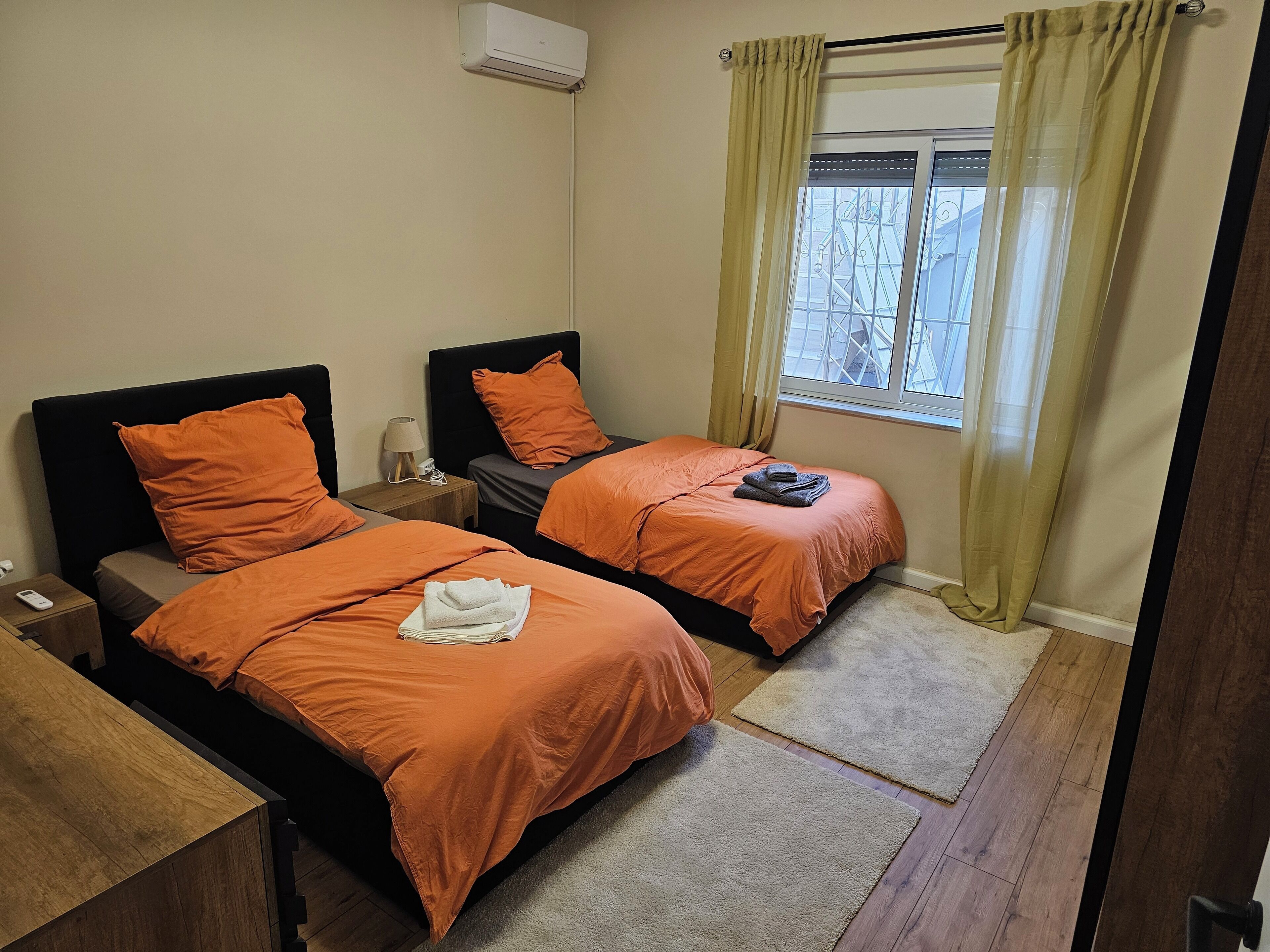 Un dormitorio con dos camas, cada una con ropa de cama naranja, una mesita de noche con lámpara y una ventana con cortinas.