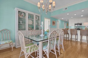 A dining area with a glass table surrounded by wicker chairs, a white cabinet with glass doors, and a kitchen in the background. - 3BR/3BA Dune Ridge Condo with Elevator, Pool & Ocean Views (Wrightsville Beach)
