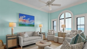 A living room with a light blue wall, a ceiling fan, a large painting, and a view of the ocean through the windows. - 3BR/3BA Dune Ridge Condo with Elevator, Pool & Ocean Views (Wrightsville Beach)