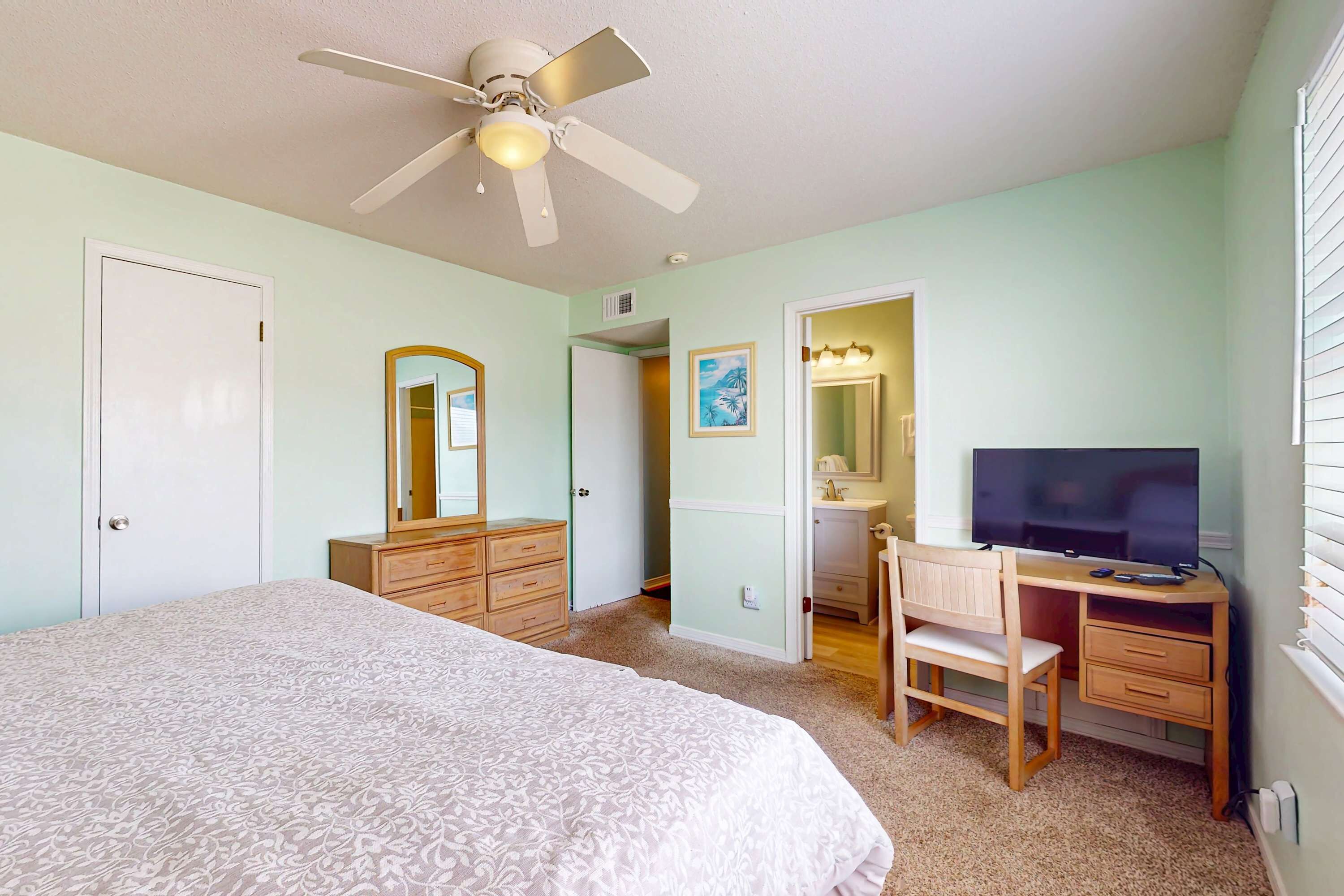 A bedroom with a bed, a desk, a chair, a television, a ceiling fan, and a door.
