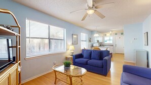 Ein Wohnzimmer mit einem blauen Sofa, einem Glastisch, einem Fernseher auf einem Schrank und einem Deckenventilator.