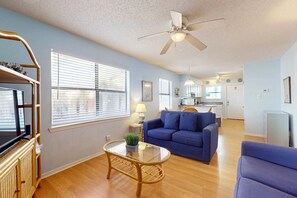 A living room with a blue sofa, a glass coffee table, a television on a stand, and a ceiling fan. - Waterfront condo with a furnished balcony, peek-a-boo Gulf views, & shared pools (Gulf Shores)
