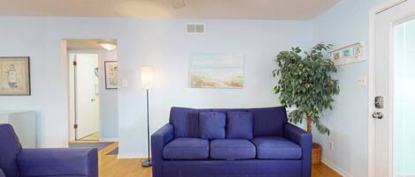 A living room with a blue sofa, a glass coffee table, a ceiling fan, and a large plant.