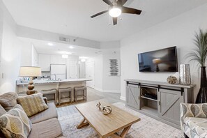 A modern living room with a sofa, coffee table, and TV. There is a kitchen area with white cabinets and a dining table. - Gulf-Front Condo w Beach Access, WiFi, Balcony & Shared Pools, Hot Tubs & Gym (Gulf Shores)