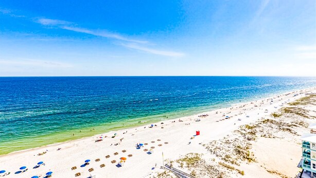 Uma praia com águas azul-claras, areal arenoso e um edifício com várias varandas.