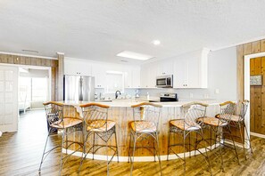 A modern kitchen with white cabinetry, a central island with bar stools, stainless steel appliances, and a wooden floor. - Beachfront house with balcony, deck, ocean view, central AC, & washer/dryer (Gulf Shores)