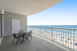 A balcony with a table and chairs, a door, and a view of a beach with umbrellas. - 6th-floor condo with pool, central AC, free WiFi, balcony, & views (Gulf Shores)