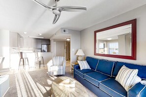 A modern living room with a blue sofa, a striped armchair, a glass coffee table, and a ceiling fan. - Lagoon-front, 3rd-floor condo with balcony, AC, & outdoor pool (Gulf Shores)