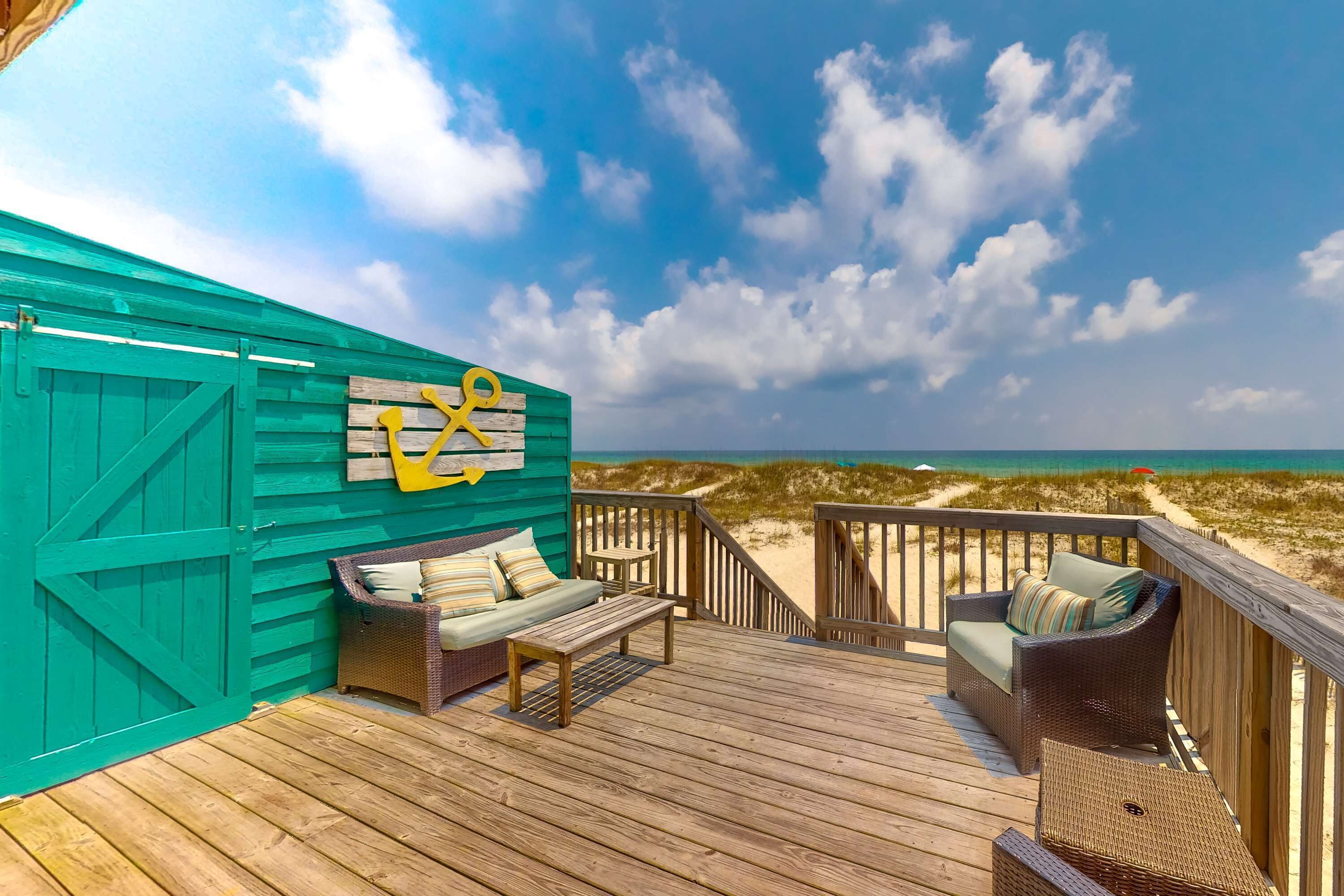 Una terraza de madera con mobiliario exterior, una pared verde con un símbolo de ancla amarillo, y vista a la playa.