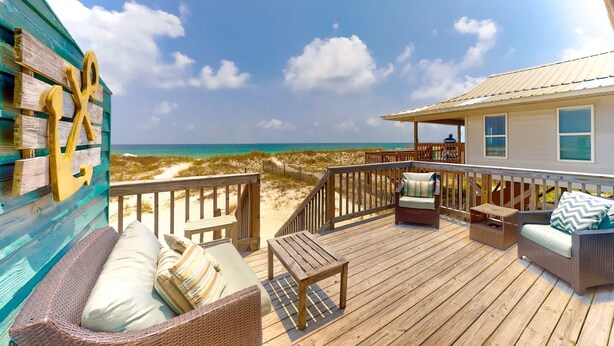 A wooden deck with outdoor furniture, a view of the beach, and a building with a metal roof. - Beachfront home with furnished waterfront deck, balcony, & central AC (Gulf Shores)