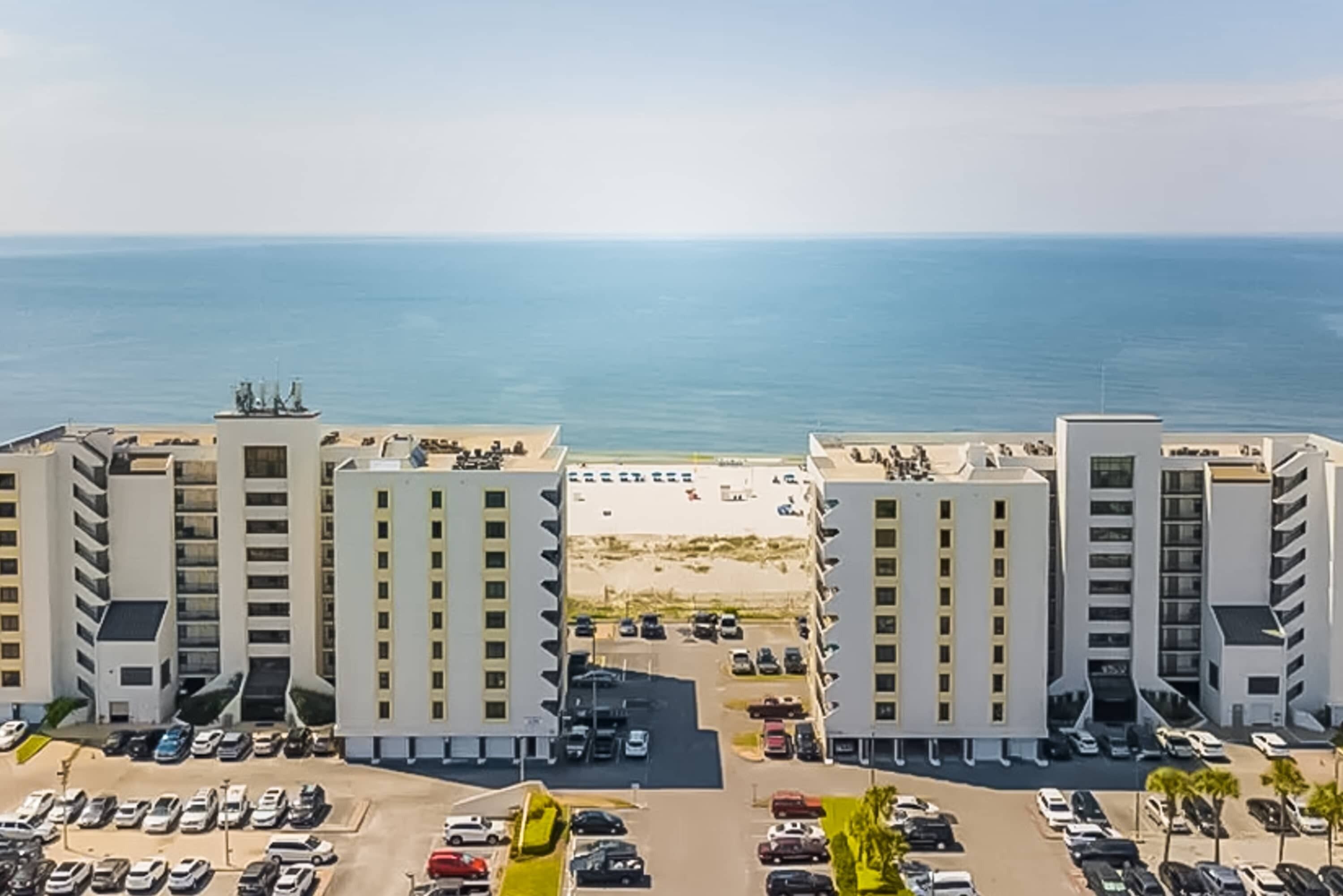 A high-rise hotel with multiple floors, a parking lot, and a beach view.