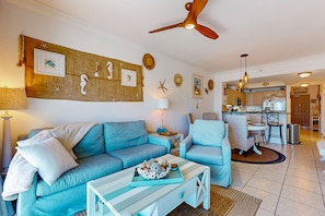 A living room with a blue sofa, a coffee table, and a dining area with a table and chairs. - Gulfview condo with skywalk to the beach, indoor & outdoor pool, & sauna (Gulf Shores)