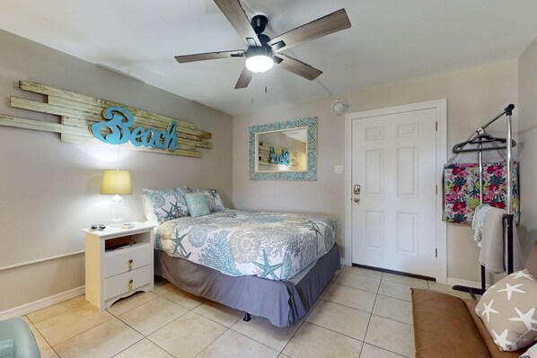 Un dormitorio con cama, mesita de noche, ventilador de techo, una puerta y una pared con un cartel que dice 'Beach'.