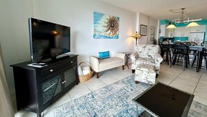 A living room with a TV, sofa, armchair, dining area, and kitchen. - 6th-floor beach-view condo with outdoor pool, tennis court, sauna, hot tub, gym (Gulf Shores)