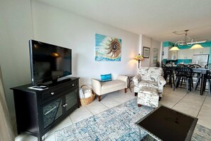 A living room with a TV, sofa, armchair, dining area, and kitchen. - 6th-floor beach-view condo with outdoor pool, tennis court, sauna, hot tub, gym (Gulf Shores)