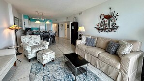 A living room with a sofa, ottoman, coffee table, and chairs. There is a dining area with a table and chairs in the background. - 6th-floor beach-view condo with outdoor pool, tennis court, sauna, hot tub, gym (Gulf Shores)
