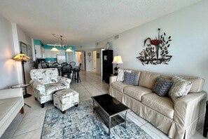 A living room with a sofa, ottoman, coffee table, and chairs. There is a dining area with a table and chairs in the background. - 6th-floor beach-view condo with outdoor pool, tennis court, sauna, hot tub, gym (Gulf Shores)