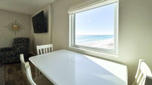 A room with a white table, a window with a beach view, a patterned chair, and a sun decoration on the wall. - Oceanfront condo with pool, grilling area, alfresco dining, internet, W/D, & AC (Gulf Shores)