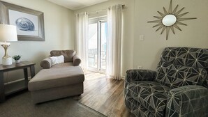 A living room with a sofa, a patterned armchair, a side table with a lamp, and a framed artwork on the wall. - Oceanfront condo with pool, grilling area, alfresco dining, internet, W/D, & AC (Gulf Shores)