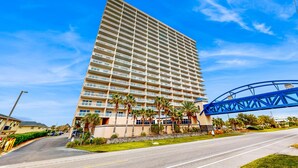 A tall hotel with multiple balconies, a pedestrian bridge, and a well-maintained lawn. - Gulf view condo with resort amenities, scenic balcony, open layout, W&D, & AC (Gulf Shores)