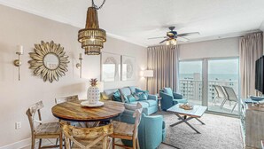 A modern living room with a round wooden dining table, blue sofa, and a balcony with a view. - Gulf view condo with resort amenities, scenic balcony, open layout, W&D, & AC (Gulf Shores)