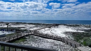 Vista costera con un paseo marítimo de madera que conduce a la playa, cielo despejado y el océano al fondo.