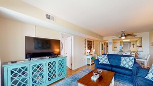 A living room with a TV, a blue sofa, a wooden coffee table, and a teal cabinet. - Inviting condo with ocean view, on-site pool, cozy balcony, washer/dryer & AC (Gulf Shores)