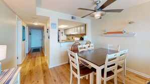 A dining area with a wooden table and white chairs, a ceiling fan, and a kitchen in the background. - Inviting condo with ocean view, on-site pool, cozy balcony, washer/dryer & AC (Gulf Shores)