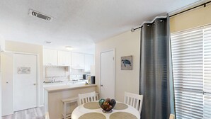 Een eetgedeelte met een ronde tafel, witte stoelen, een keuken op de achtergrond en een raam met zonwering.