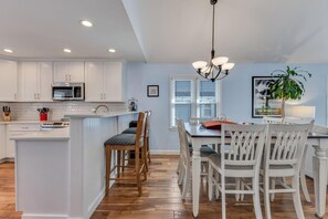 A modern kitchen with white cabinetry, a central island, and a dining area with white chairs and a wooden table. - Kraken - Spacious 4 Bedroom Home with Private Beach Access (Kure Beach)