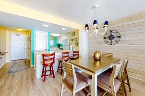 A dining area with a wooden table and chairs, a chandelier, and a compass wall decoration. - Fourth-floor Gulf-front getaway with balcony, views, central AC, & outdoor pool (Gulf Shores)