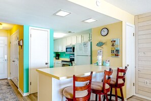 A kitchen with white cabinets, a microwave, and a refrigerator. There is a clock on the wall and a small fish decoration. - Fourth-floor Gulf-front getaway with balcony, views, central AC, & outdoor pool (Gulf Shores)