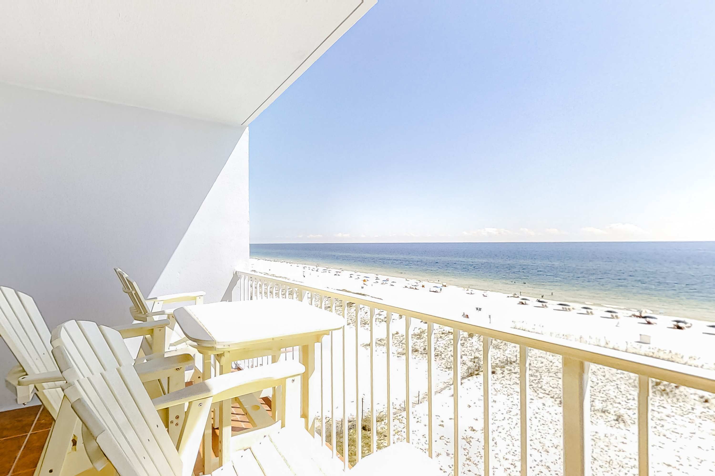 Ein Balkon mit weißen Stühlen und einem Tisch, mit Blick auf einen Strand mit Sonnenschirmen.
