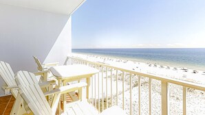 Varanda com cadeiras e mesa brancas, com vista para uma praia com guarda-sóis.