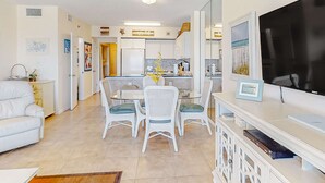 Uma sala de estar moderna com um sofá, mesa de jantar com cadeiras, televisão e área de cozinha ao fundo.