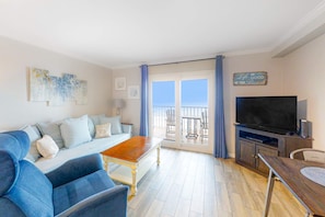 A living room with a sofa, coffee table, TV, and balcony with outdoor furniture. - Attractive and Convenient Gulf-Front Home Right on Beach with Shared Pool, Hot Tub (Gulf Shores)