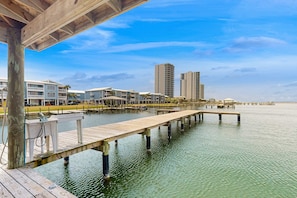 A waterfront property with a wooden dock extending into the water, surrounded by modern buildings and a clear sky. - Cheerful Condo with Shared Pool, Pier, and Beach Access (Gulf Shores)