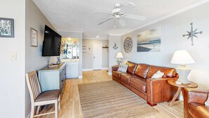 Een woonkamer met een bank, een stoel, een televisie, een plafondventilator en een keuken op de achtergrond.