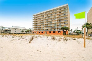 Ein Strandhotel mit mehreren Balkonen, ein Sandstrand davor und eine grüne Flagge.