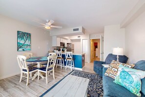 Ein modernes Wohnzimmer mit einem blauen Sofa, einem Esstisch mit Stühlen, einer Küchenzeile mit blauer Kochinsel und einem Deckenventilator.
