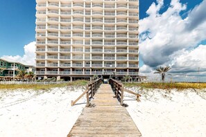 Ein Hotel direkt am Strand mit einer Holzpromenade, die zum Eingang führt, mehreren Balkonen und blauem Himmel.
