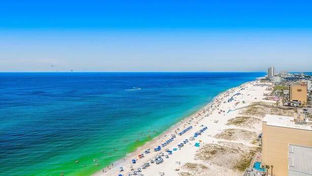 Une plage avec une eau d’un bleu limpide, un rivage sablonneux et des bâtiments en arrière-plan.