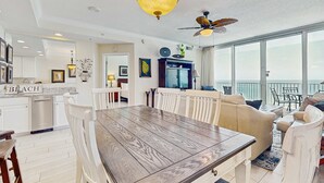 Un vaste espace de vie comprenant une table à manger, des chaises blanches, un ventilateur de plafond et une grande fenêtre donnant sur un paysage agréable.