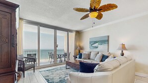 Un vaste espace de vie comprenant un canapé blanc, une table à manger et des chaises, un ventilateur de plafond et une grande fenêtre donnant sur l’océan.