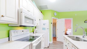 Une cuisine avec des appareils électroménagers blancs, une horloge au mur et une porte donnant sur une autre pièce.