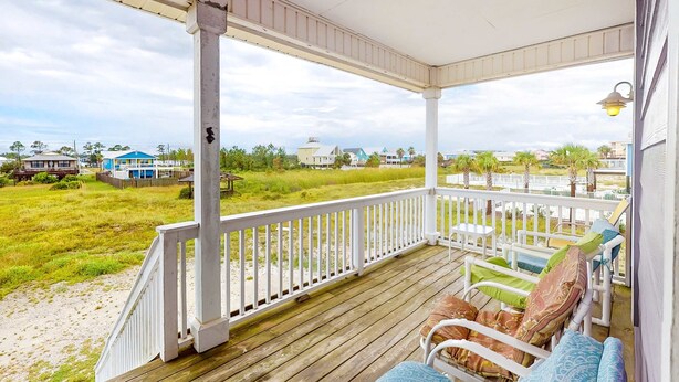 Une véranda couverte avec une balustrade blanche, une terrasse en bois et une vue sur la plage et les maisons au loin.