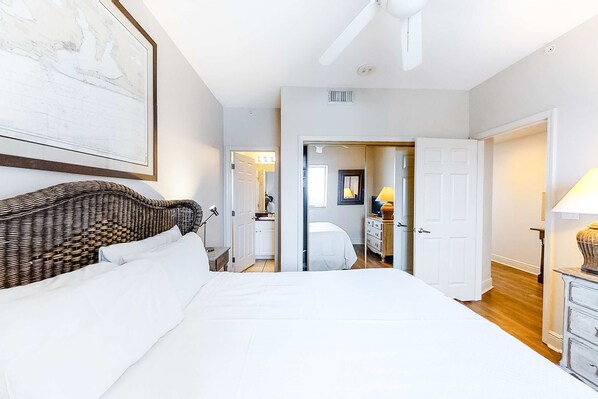 A bedroom with a large bed, a wicker headboard, a ceiling fan, and a door leading to a bathroom. - Gulf-front condo with pool, sauna, washer dryer, central AC, & gym (Gulf Shores)