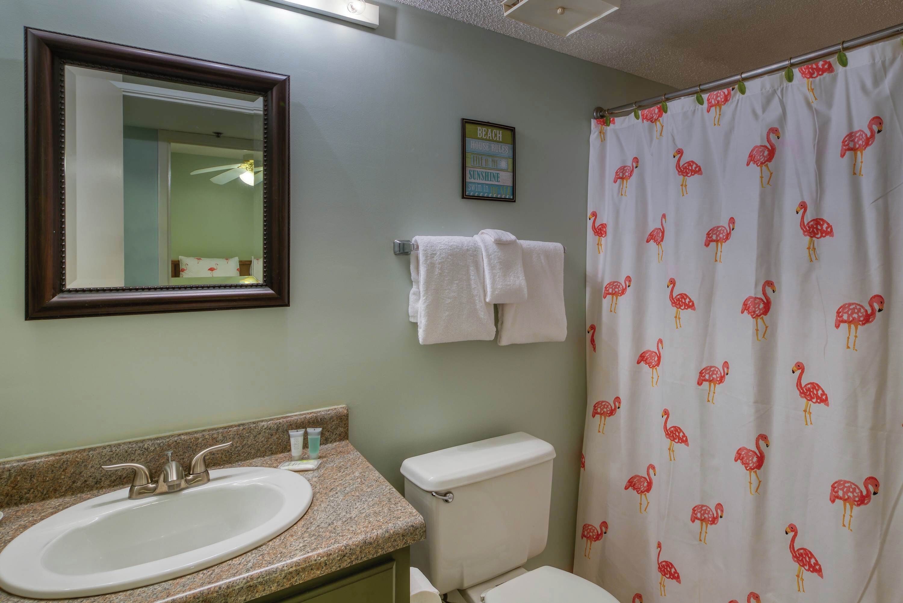 A bathroom with a sink, mirror, toilet, and shower curtain with a flamingo pattern.