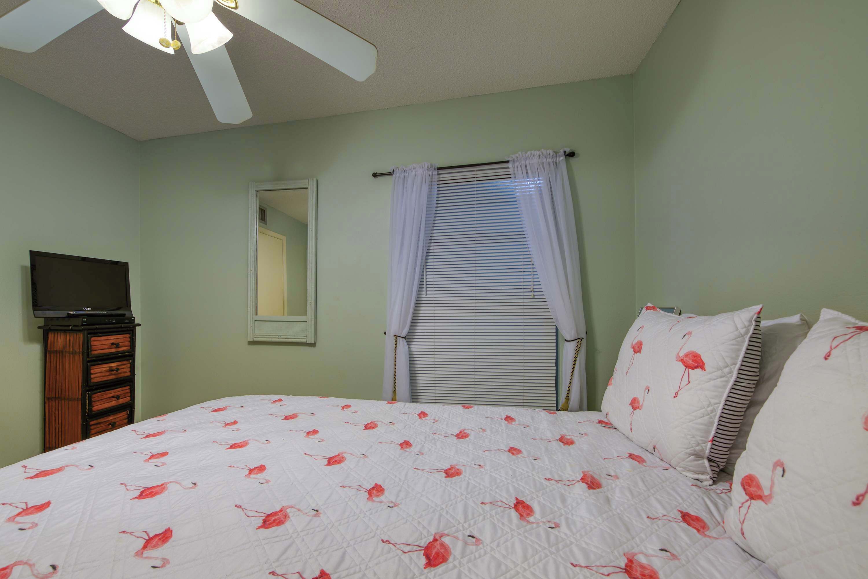 A bedroom with a bed, a television on a stand, a window with curtains, and a ceiling fan.